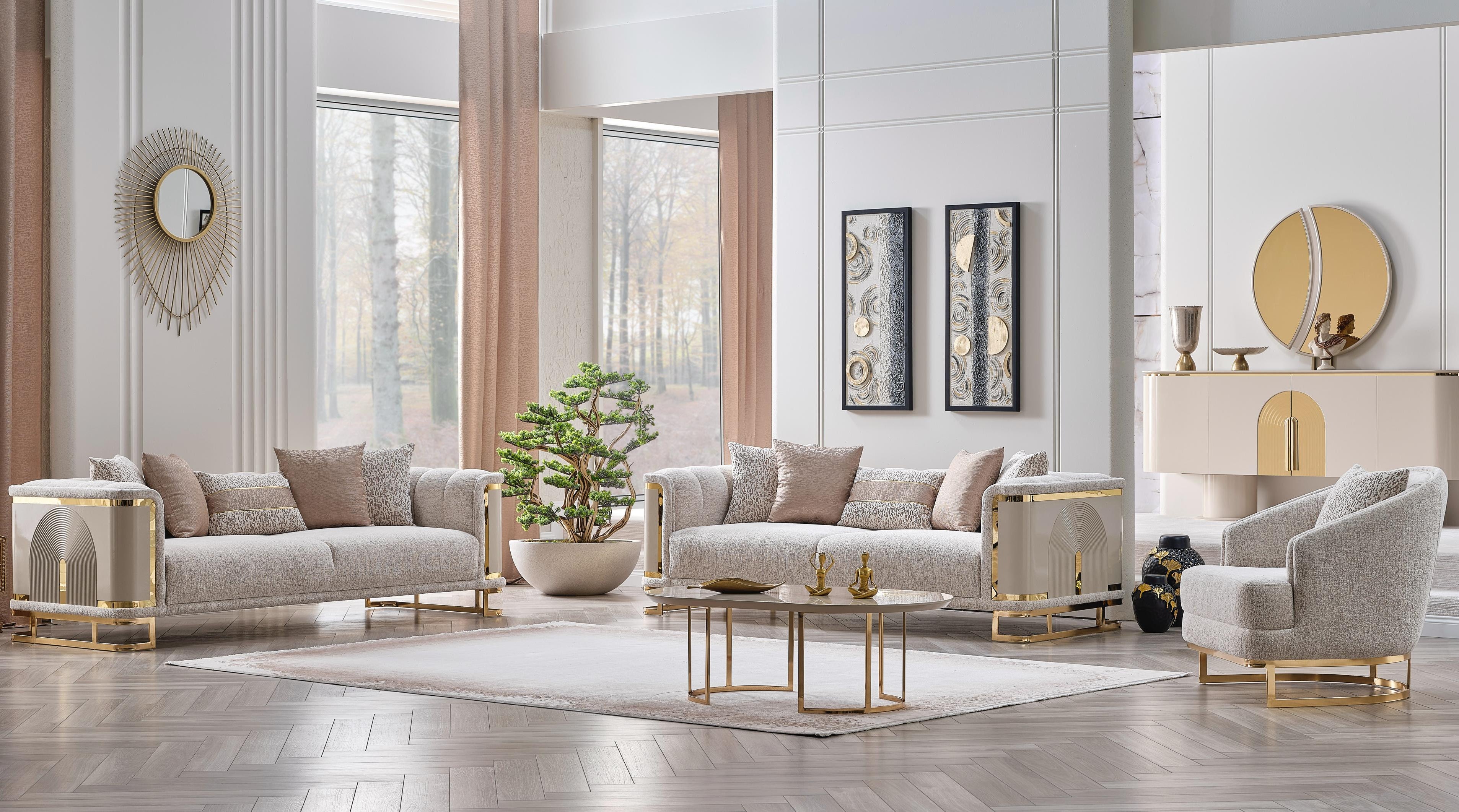 A luxury modern living room set featuring a light beige sofa, matching armchair, and a small round coffee table with gold metal legs, arranged in a bright, elegant room with beige walls, wood floors, and decorative accents.