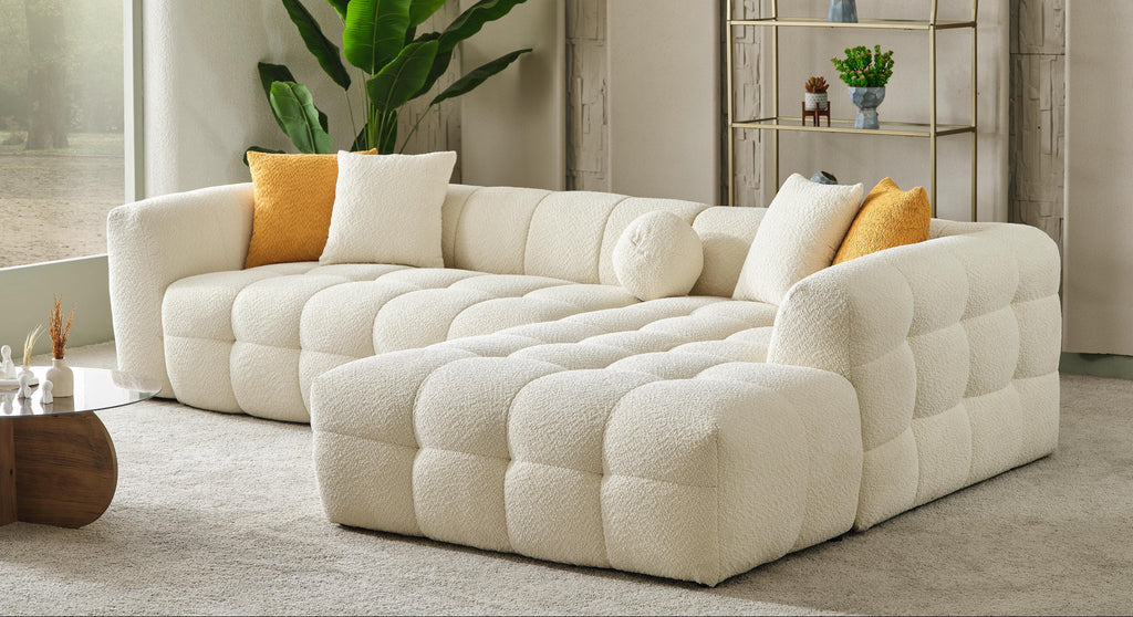 A modern tufted sectional sofa in cream bouclé fabric, styled in a bright, contemporary living room with yellow and white accent pillows, a large green plant, and a gold-framed shelving unit.