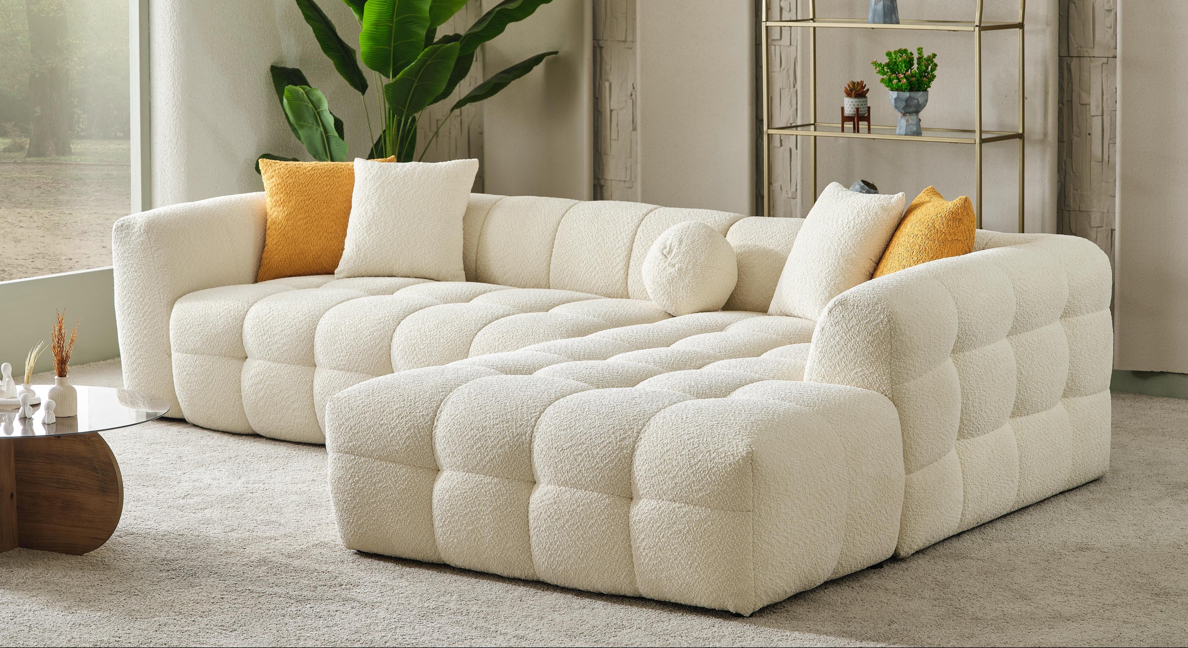 A modern tufted sectional sofa in cream bouclé fabric, styled in a bright, contemporary living room with yellow and white accent pillows, a large green plant, and a gold-framed shelving unit.